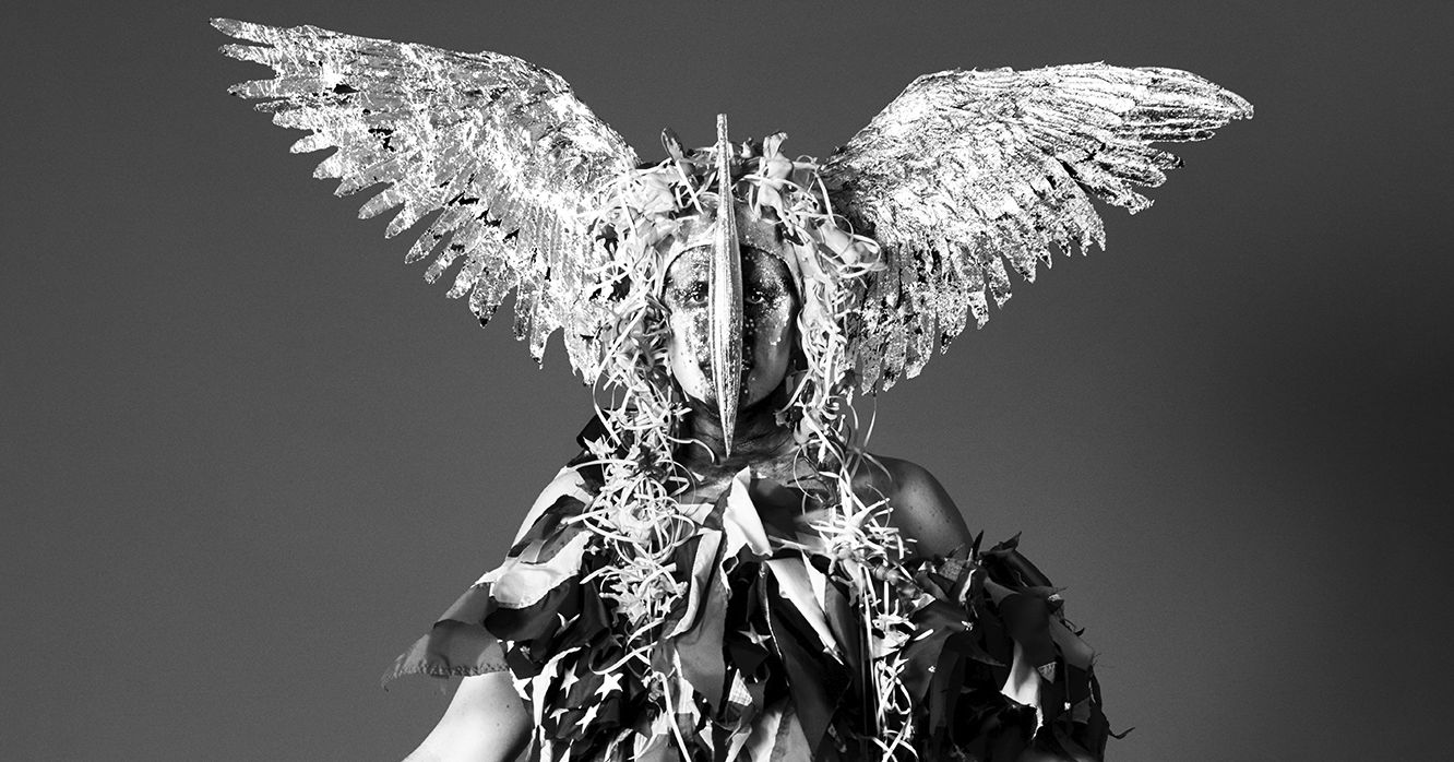 A person stares ahead. His elaborate costume includes a winged headpiece that obscures his face and a garment made from tatters of the United States flag.
